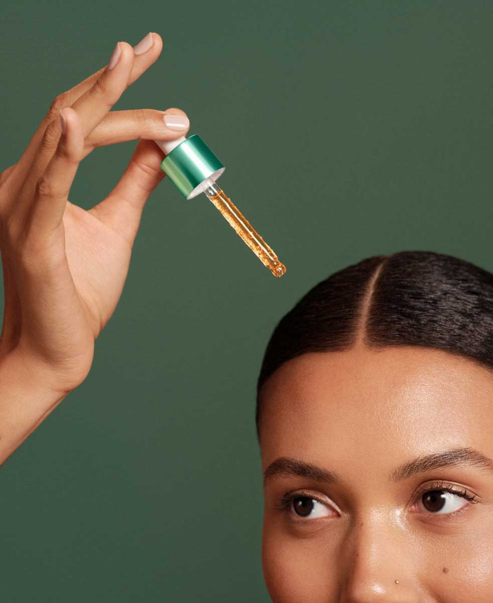 Woman with thick, curly hair in a low bun holding the serum dropper over her head in front of a dark green background. 