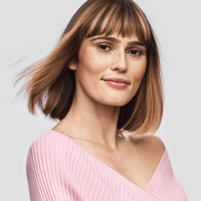 Woman with short blonde hair wearing a pink sweater in front of a grey background. 