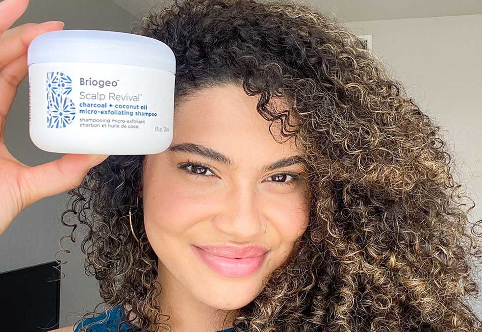 Woman with very curly, brown hair holding the Scalp Revival Exfoliating Scrub by her face. 