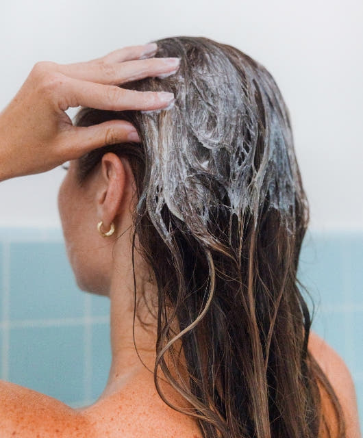 A woman with brown hair washing her scalp with Briogeo scalp scrub in a blue tiled shower. 